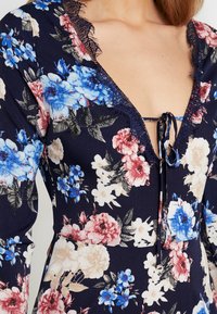 Mönstrad klänning i marinblått med blommor i blått, rosa och vitt. Designen har en djup V-ringning, spetsdetaljer och knytning fram.
