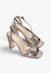 Et par metallic sølv høje hæle sandaler med firkantede tæer, ankelremme og åbne sider vist på en hvid baggrund.