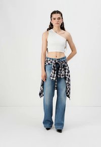 Witte een-schouder crop top, blauwe wijde jeans en een geruit flanellen shirt om de taille geknoopt. Zwarte hoge hakken maken de look compleet.