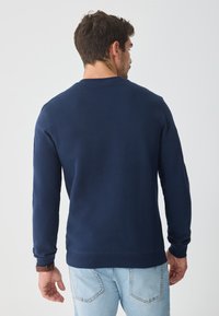 Námornícky modré mikina vyrobená z jemného materiálu, s dlhými rukávmi, okrúhlym výstrihom a rebrovaným lemom. Zobrazená zozadu s svetlými džínsami.