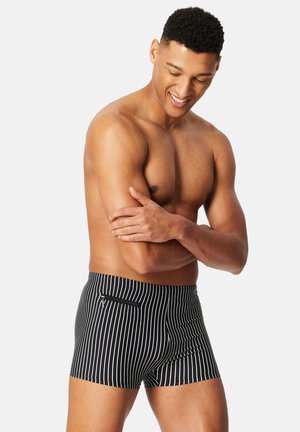 Homme musclé portant un maillot de bain noir à rayures verticales blanches et une poche zippée, souriant et regardant vers le bas, les bras croisés.