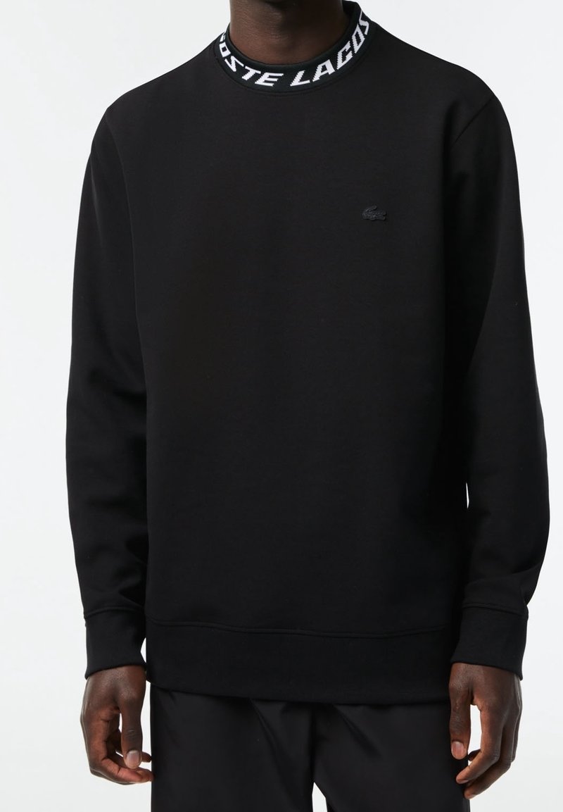 Homme portant un sweat-shirt noir avec le texte blanc « LACOSTE » sur le col et un petit logo crocodile sur la poitrine gauche.