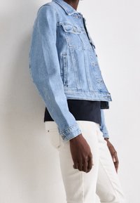 Ljusblå denimjacka med klassisk skärning, knappstängning, bröstfickor och något slitna kanter. Bärs över en mörk topp och ljusa byxor.