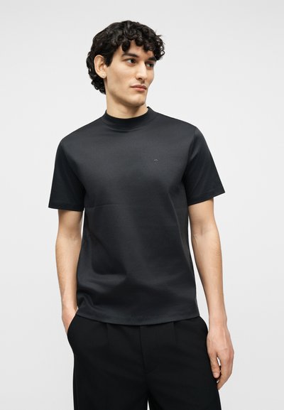 Άνδρας με σγουρά μαλλιά που φοράει απλό μαύρο κοντομάνικο πουκάμισο με ψεύτικο γιακά και μαύρο παντελόνι, με τα χέρια στις τσέπες, κοιτάζοντας δεξιά.