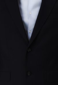 Navy blazer featuring a two-button closure, notched lapels, and side pockets. Underneath, a light blue dress shirt with a classic collar and buttons.