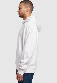 Weißer Kapuzenpullover aus weichem Material, mit einer vorderen Tasche und gerippten Bündchen. Im Profil mit blauen Jeans getragen.