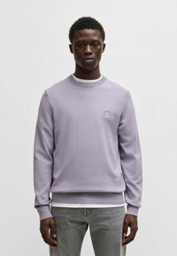 Helles lila Strick-Sweatshirt mit rundem Ausschnitt, gerippten Bündchen und Saum. Verfügt über ein kleines quadratisches Logo auf der Brust. Wird über einem weißen Hemd getragen.