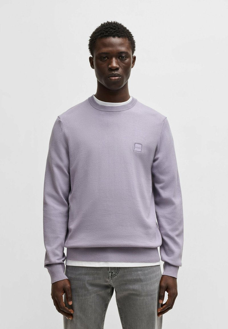 Helles lila Strick-Sweatshirt mit rundem Ausschnitt, gerippten Bündchen und Saum. Verfügt über ein kleines quadratisches Logo auf der Brust. Wird über einem weißen Hemd getragen.