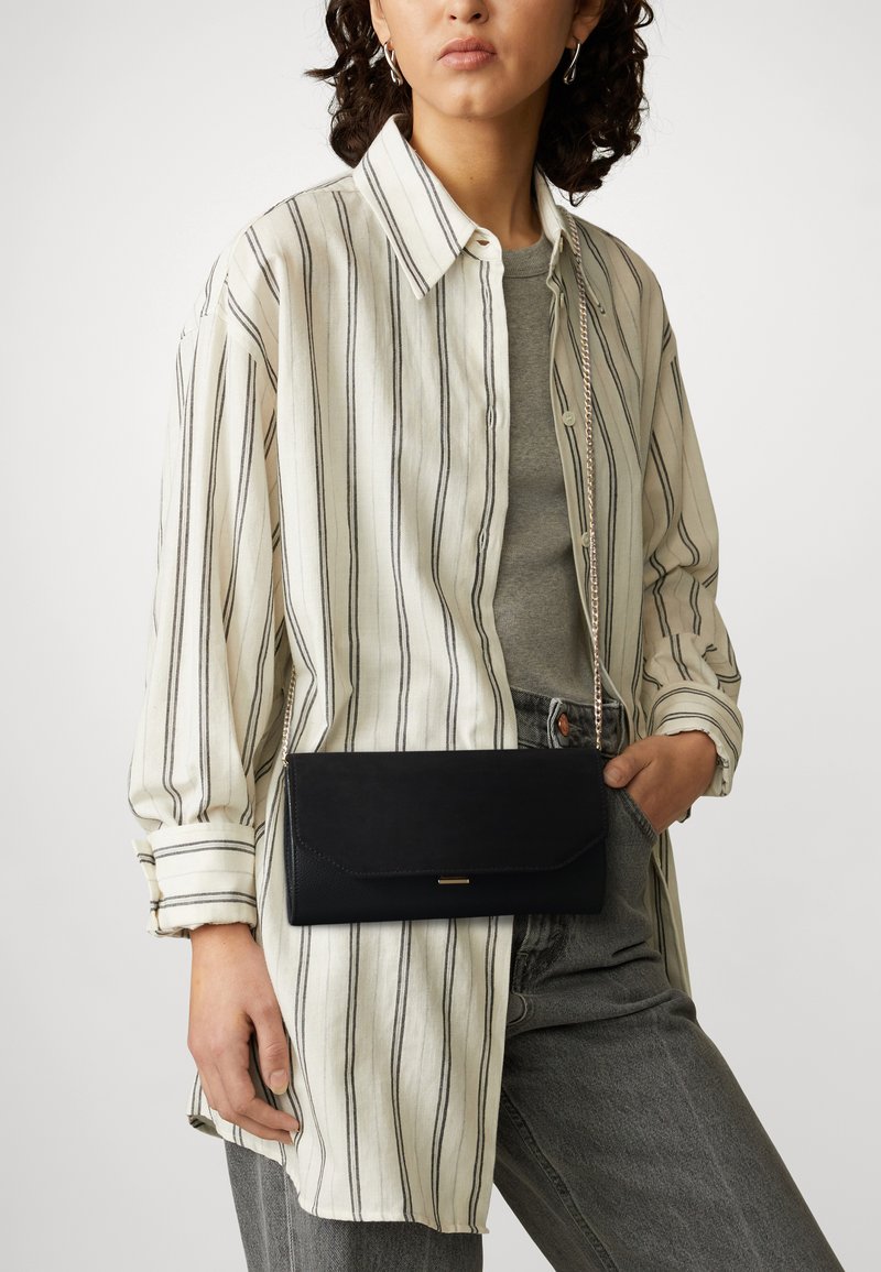 Black leather clutch with a fold-over top, chain strap, and a metallic accent. Worn with a striped shirt and grey top, paired with light jeans.