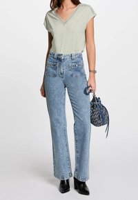 Jasnozielona bluzka z dekoltem w serek i koronkowym wykończeniem; wysokie, jasnoniebieskie dżinsy; czarne botki na obcasie; wzorzysta torebka w niebiesko-białe wzory.