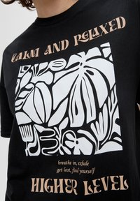 Schwarzes Baumwoll-T-Shirt mit einem großen weißen abstrakten Pflanzendesign. Der Text in hellbraun lautet "RUHIG UND ENTSPANNT", mit kleinerem Text darunter.
