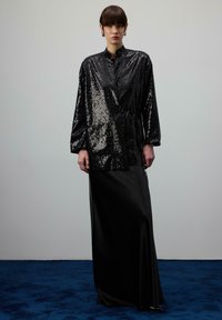 Osoba v dlhom čiernom saténovom sukni a čiernom flitrovanom saku so stojatým golierom stojaca na modrom koberci pred sivou stenou.