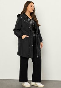 Impermeabile nero con cappuccio, polsini elastici e vita con cordoncino. Indossato sopra una camicia a righe bianche e nere e abbinato a pantaloni neri a zampa d'elefante e sneakers bianche.