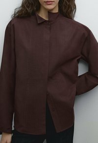 Chemise à manches longues bordeaux avec un col mandarins, présentant des détails de plis verticaux et une fermeture à boutons sur le devant. Texture du tissu lisse.