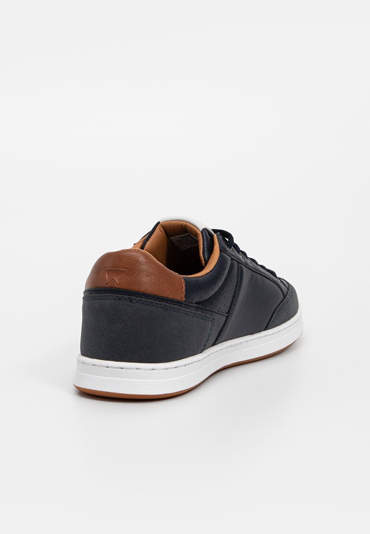 Zwarte leren sneaker met een bruine leren hakaccent, witte zool en geperforeerde details aan de zijkanten. Ronde neus en vetersluiting.