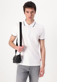 Balta polo marškiniai su juodu apykakle ir rankovių akcentais, derinami su šviesiai pilkais džinsais. Modelis laiko mažą juodą kryžminį krepšį.