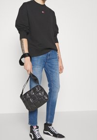 Svart sweatshirt med logotyp, blå jeans, svart quiltad handväska och svarta högskaftade sneakers med vita detaljer. Detaljer av avslappnad outfit synliga.