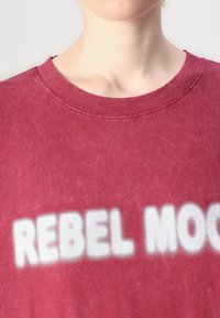 Μπορντό βαμβακερό t-shirt με στρογγυλή λαιμόκοψη και λευκό κείμενο "REBEL MOON" εκτυπωμένο σε έντονο, απαλό εστιασμένο στυλ πάνω από το στήθος.