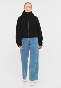 Giacca in pile nera con collo alto, chiusura con zip e due tasche frontali, abbinata a jeans a gamba larga blu chiaro e scarpe da ginnastica bianche.