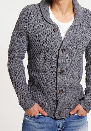 Hombre con cárdigan de punto gris texturizado con botones de madera sobre camisa blanca, con las manos en los bolsillos de los vaqueros azules.