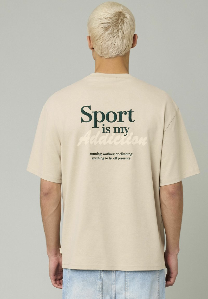 T-shirt en coton beige avec du texte vert au dos : "Le sport est ma dépendance" avec un texte supplémentaire en dessous. Encolure classique et coupe décontractée.