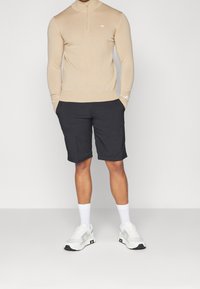 Beige ziptröja, svarta shorts och vita strumpor med grå sneakers. Outfiten har en åtsittande design och minimal logotyp.