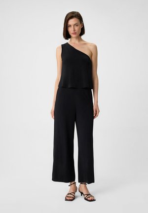 Zwarte jumpsuit met asymmetrisch ontwerp, voorzien van een mouwloze top en wijde pijpen. Soepele textuur en minimalistische stijl. Strappy zwarte sandalen.