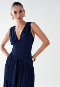 Marineblaues Kleid aus strukturiertem Stoff; verfügt über einen tiefen V-Ausschnitt, vertikale Falten und ein asymmetrisches Design mit ausgestelltem Rock. Goldene, runde Ohrringe.