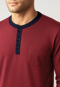 Camicia bordeaux a maniche lunghe con colletto blu navy, patta con tre bottoni e una tasca sul petto. Realizzata in tessuto liscio e morbido.