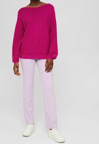 Fuksjowy oversize sweter z długimi rękawami, połączony z jasnofioletowymi prostymi spodniami i białymi trampkami, na modelce na jednolitym tle.