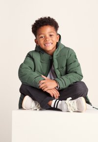 Green padded jacket with a hood, gray t-shirt, dark jeans, and gray sneakers. The jacket features quilted texture and a front zipper.