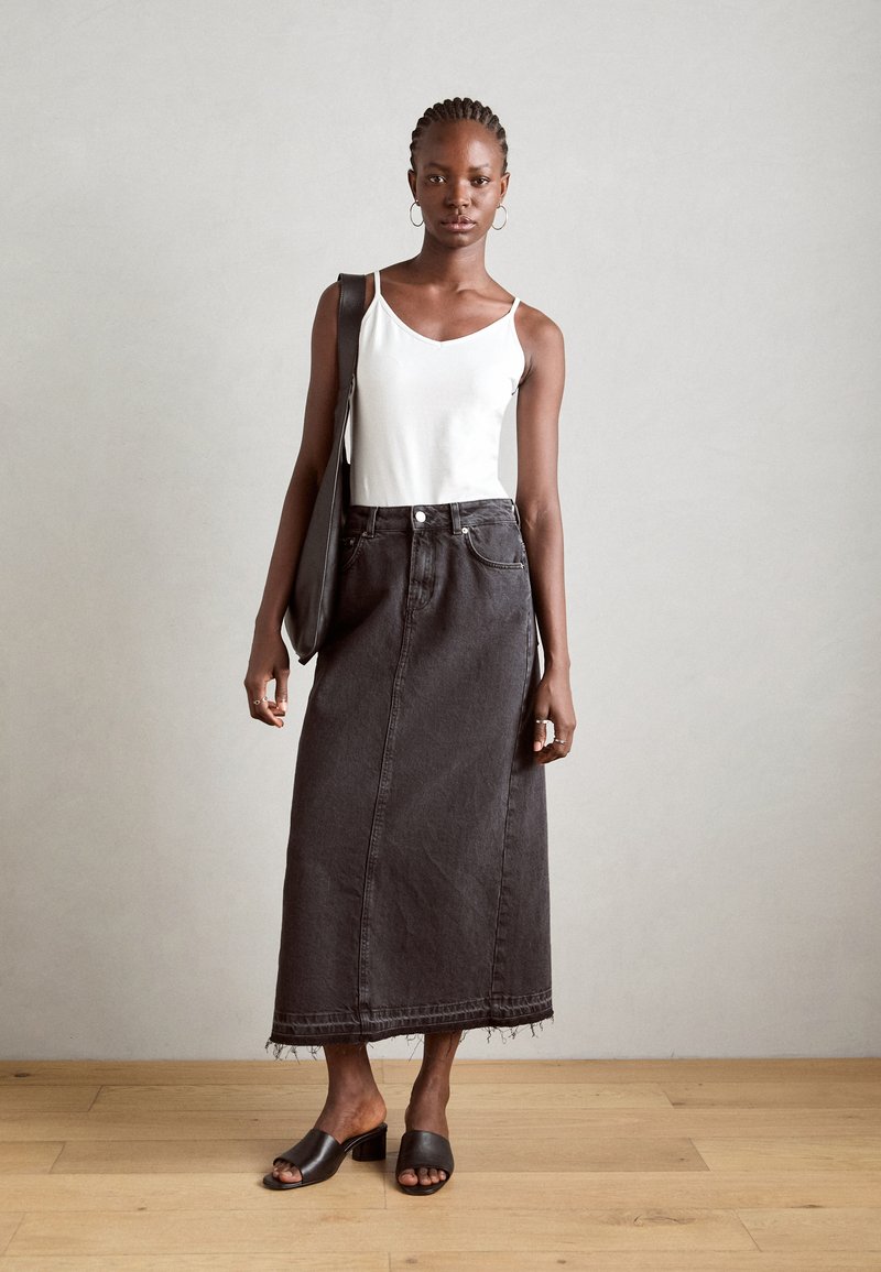 Zwarte denim maxi-rok met een rafelige zoom, gecombineerd met een witte mouwloze top en zwarte hoge muiltjes; model met een zwarte handtas.