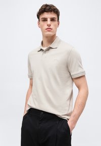 Jeune homme portant un polo beige clair avec le logo "BOSS" et un pantalon noir, debout les mains dans les poches devant un fond uni.