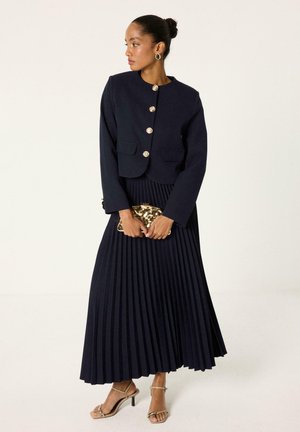 Femme portant un blazer bleu marine avec des boutons dorés et une jupe maxi plissée assortie, tenant une pochette dorée, debout devant un fond neutre.
