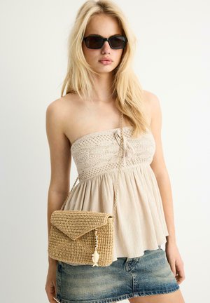 Plavolaska ženska z rjavimi sončnimi očali, brez naramnic bež topom, jeans miniskirt in tkanim rjavim torbico čez ramo z obeskom v obliki ribice iz perlic.