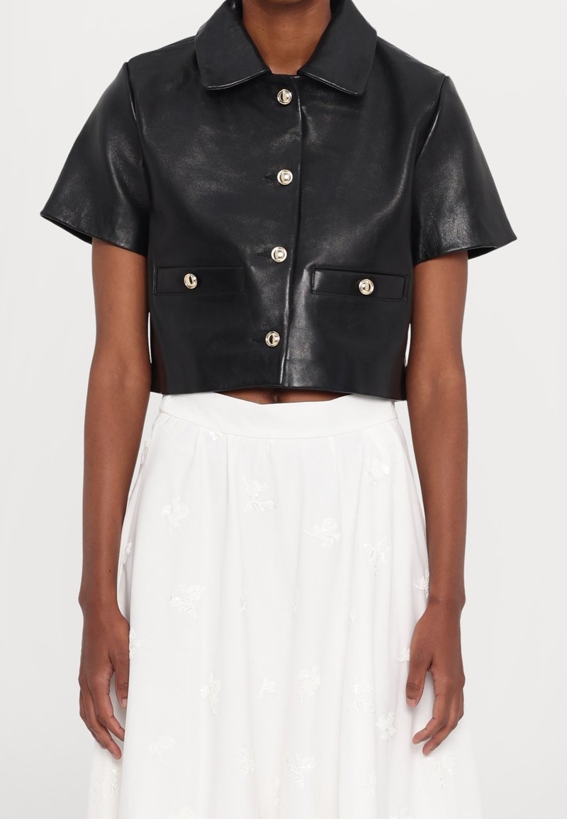 Veste courte noire à manches courtes en similicuir avec cinq boutons sur le devant, portée sur une jupe blanche taille haute avec une broderie florale subtile.