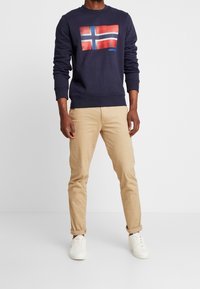 Felpa navy con grafica della bandiera norvegese, abbinata a pantaloni beige chiaro e sneakers bianche. Semplice colletto a giro e polsini a coste.
