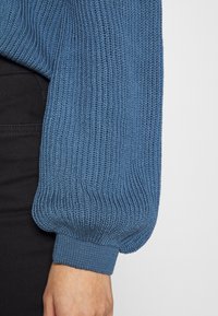Gebreide mouw van een sweater in een zachte blauwe kleur met ribbeltextuur en een aangesloten manchet. Het materiaal lijkt lichtgewicht en rekbaar.