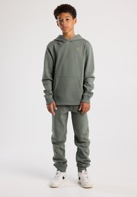Grüne Kapuzenjacke und passende Jogginghosen aus weichem, strukturiertem Stoff. Mit einer Fronttasche und gerippten Bündchen. Getragen mit weißen Sneakers.