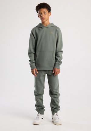 Groene hoodie en bijpassende joggers van zachte, gestructureerde stof. Heeft een voorkantzak en ribgebreide manchetten. Gekenmerkt met witte sneakers.