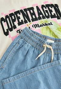 Hvid t-shirt med sort "COPENHAGEN" tekst og lyserød ternet baggrund, sammen med blå og hvide stribede shorts med elastisk talje.