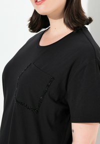 Musta puuvillapaita, jossa on pyöreä kaula-aukko ja lyhyet hihat, ja vasemmalla puolella koristeellinen jalokivielementeillä varustettu tasku.