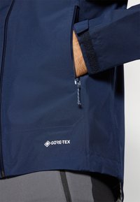 Marinblå GORE-TEX-jacka med en dragkedjeficka, kardborreband vid ärmslut och slät, vattentät tyg. Har en logotyp längst ner på fållen.
