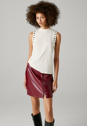 Jonge vrouw met krullend haar, draagt een mouwloze witte top met knoopdetails, een glanzende bordeauxrode minirok en zwarte kniehoge laarzen.