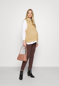 Colete de malha bege sobre uma camisa branca, calças castanhas, botas pretas de tornozelo. A segurar uma mala de mão em formato de croco rosa. Design simples e moderno.