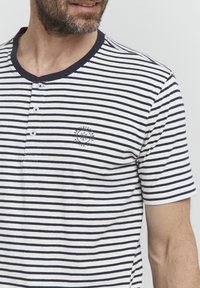 Camiseta de manga corta a rayas en blanco y azul marino, con cuello henley abotonado, textura suave y un pequeño logo en el pecho.