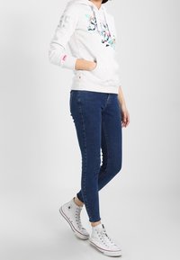 Felpa grigia chiara con design floreale, tasca frontale e coulisse, abbinata a jeans skinny blu scuro e sneaker alte bianche.
