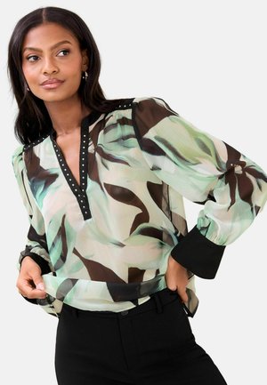 Frau trägt eine durchsichtige, gemusterte Bluse mit schwarzem Besatz und Manschetten, kombiniert mit schwarzen Hosen, posiert und richtet den Saum der Bluse mit den Händen.