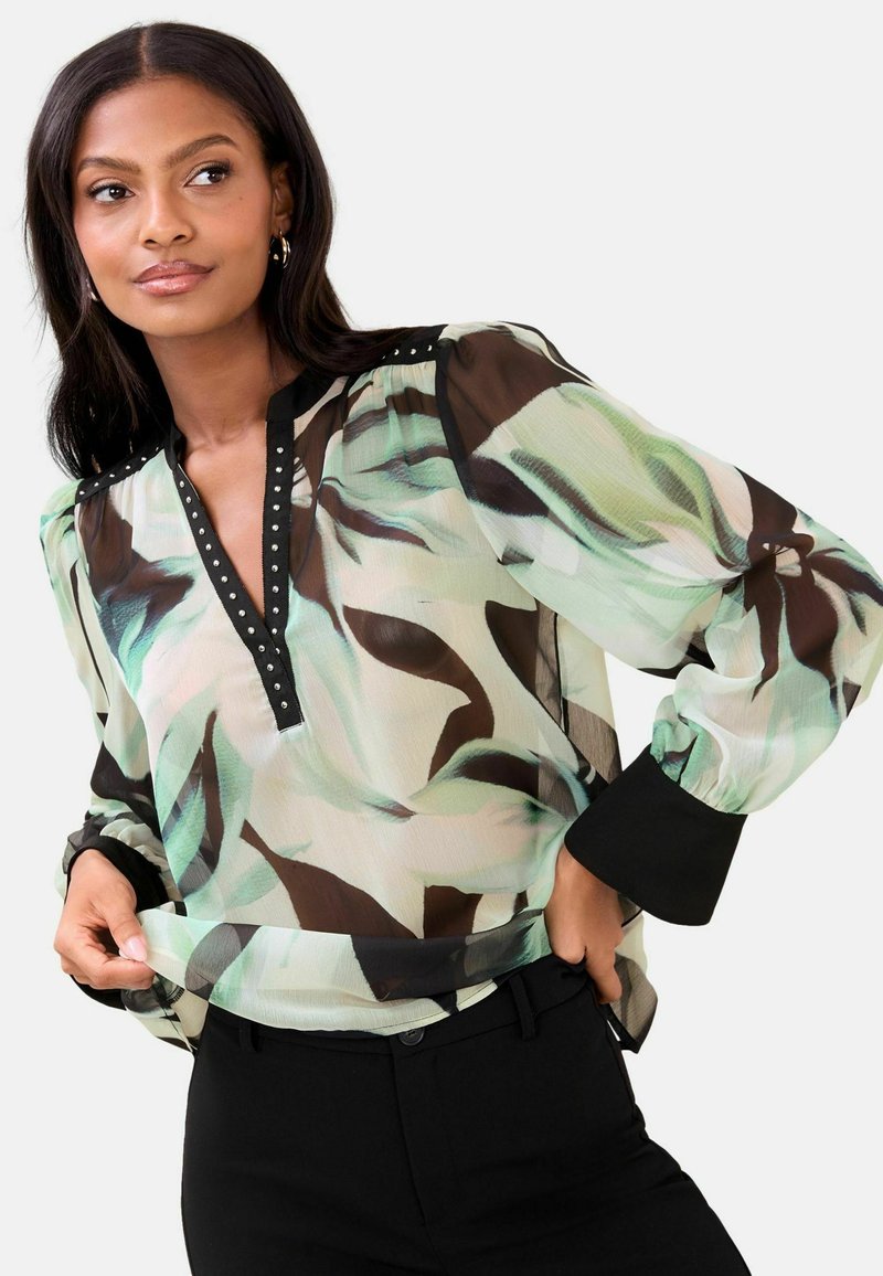 Žena v průsvitné blůze s vzorem, černým lemováním a manžetami, v černých kalhotách, pózuje s rukama upravujícími lem blůzy.