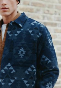 Hladná flanelová košeľa s námorníckym vzorom a geometrickými motívmi v rôznych odtieňoch modrej, s prednou vreckom a špicatým golierom.